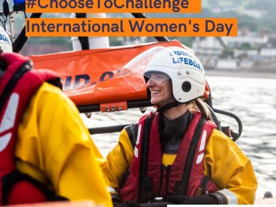 A Sidmouth Lifeboat crew member, who also happens to be female, smiles whilst onbaord the lifeboat. The text overlay reads #ChooseToChallenge International Women's Day